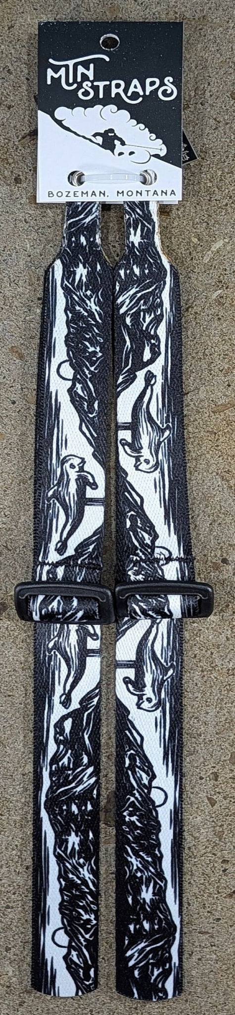 A pair of black and white patterned Mtn Straps ski pole straps with black plastic buckles, shown hanging from their original black and white cardboard header card on a textured beige background.