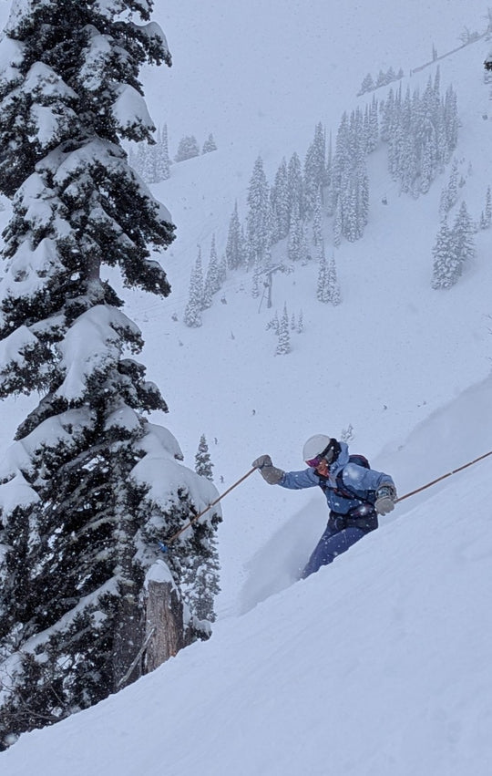 Person skiing down a snowy slope with trees on a mountain with Mtn Straps pole straps